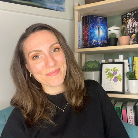 Mila Ilbach lächelt, im Hintergrund ein Bücherregal mit ihren Veröffentlichungen.