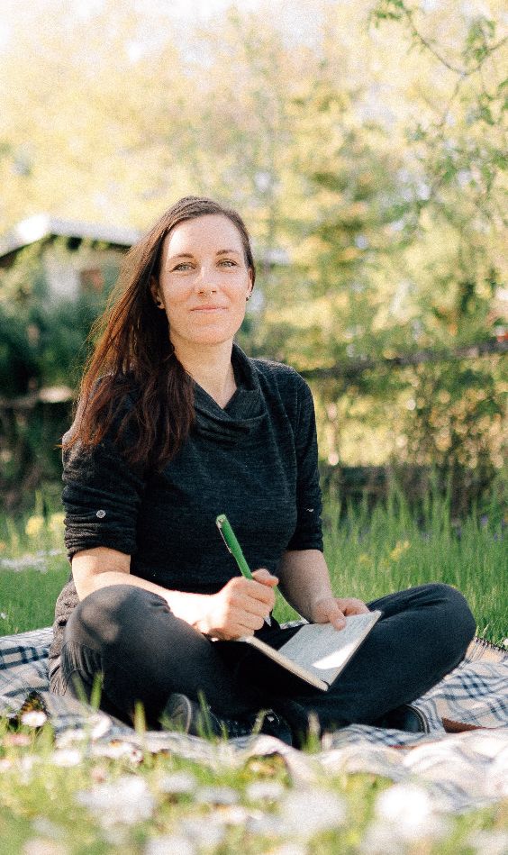 Susann sitzt auf einer Wiese, einen Stift und ein Notizbuch in der Hand.