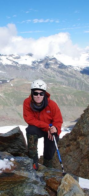 Susann in den Alpen beim Bergsteigen. Sie trägt einen Kletterhelm und eine orangefarbene Jacke. Im Hintergrund sind schneebedeckte Berge.
