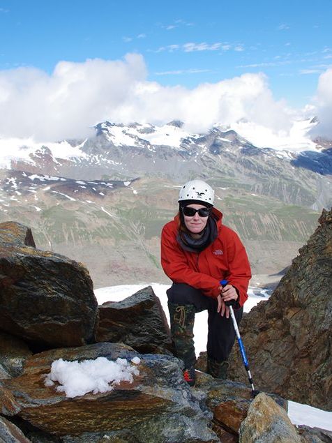 Susann in den Alpen beim Bergsteigen. Sie trägt einen Kletterhelm und eine orangefarbene Jacke. Im Hintergrund sind schneebedeckte Berge.