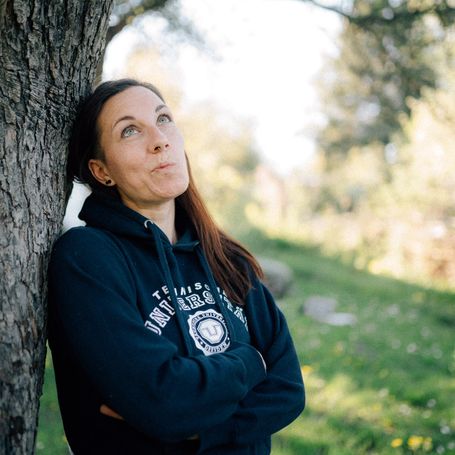 Susann in einem Hoodie an einen Baum gelegt und den Mund halb verzogen zu einem nachdenklichen Lächeln.